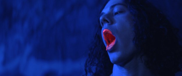 Acteur uit film Hard Paint in donkerblauw licht met neon zalmroze verf die langzaam uit zijn mond komt