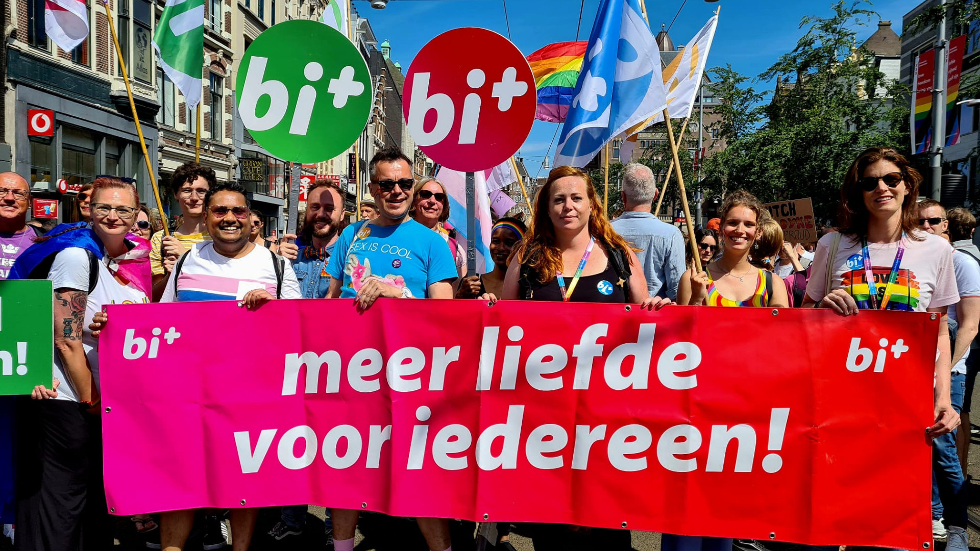 Foto van het Bi+ blok tijdens de Pride Walk. Een aantal mensen staat achter de roze banner met de tekst 'meer liefde voor iedereen!' en links- en rechtsboven het Bi+ Nederland logo.