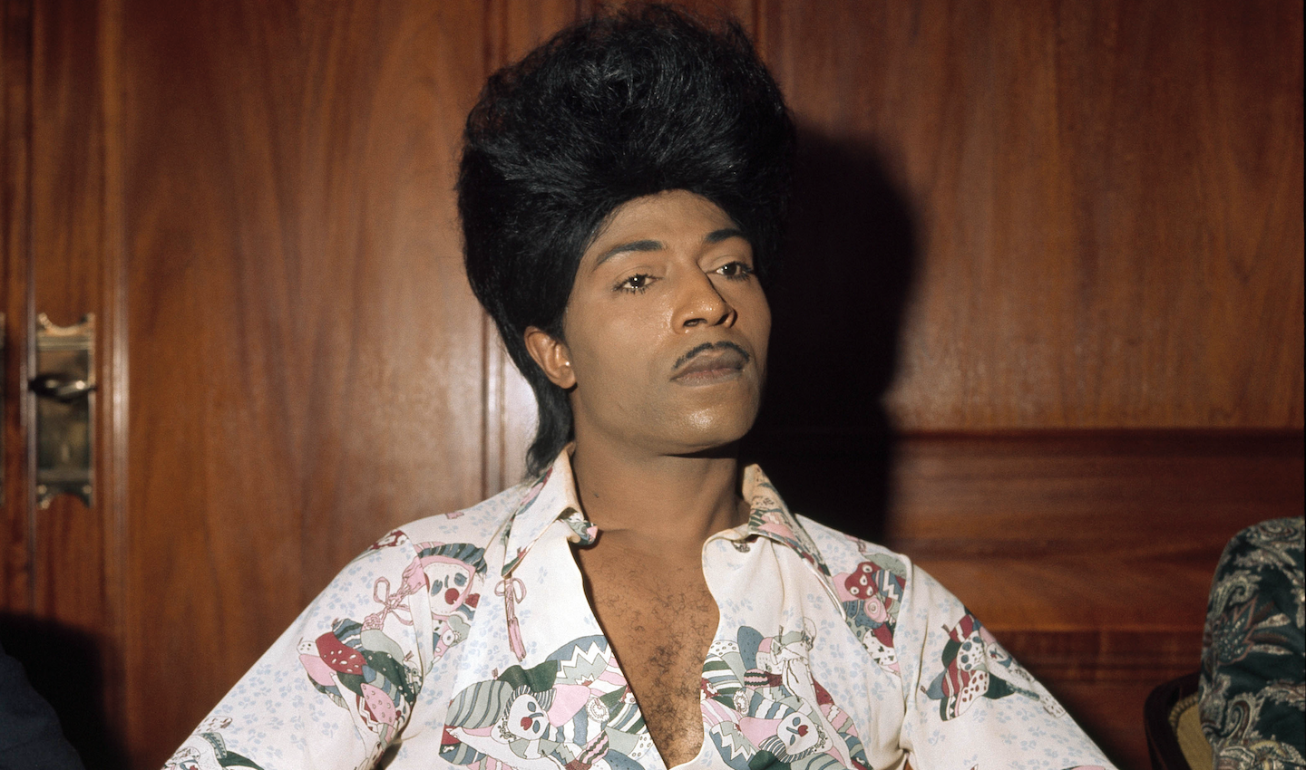 Kleurenfoto van muzikant Little Richard. Hij draagt zijn zwarte haar hoog opgestoken in pompadour-stijl. Hij heeft een witte blouse aan met grijs-roze design en kijkt geconcentreerd naar rechts. Hij zit voor een houtbruine kast..